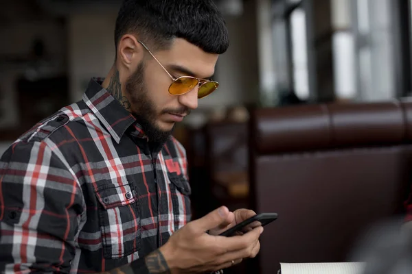 Elegante hombre con camisa a cuadros usando teléfono inteligente sentado en la cafetería —  Fotos de Stock