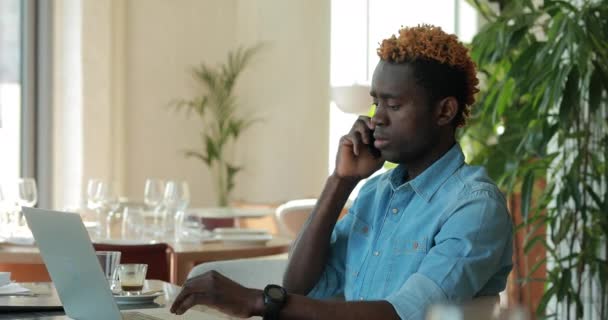 Hombre de negocios afroamericano feliz usando computadora portátil en la cafetería mientras habla por teléfono — Vídeos de Stock