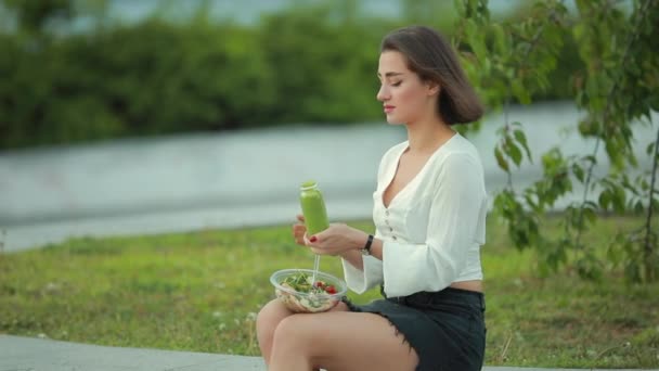 Krásná žena jedla salát, jak sedí v parku a pije hladkou — Stock video