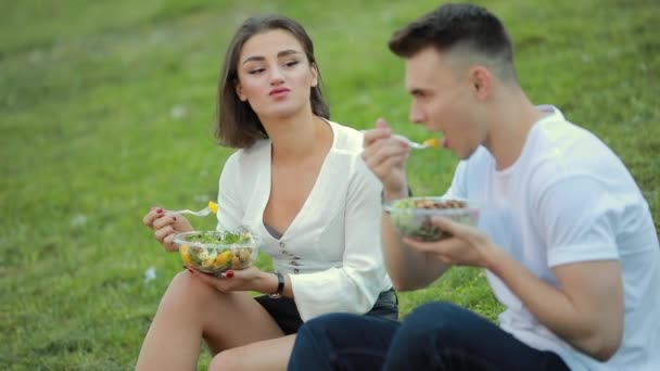 Pareja joven comiendo ensalada sentada en la hierba, picnic juntos — Vídeo de stock