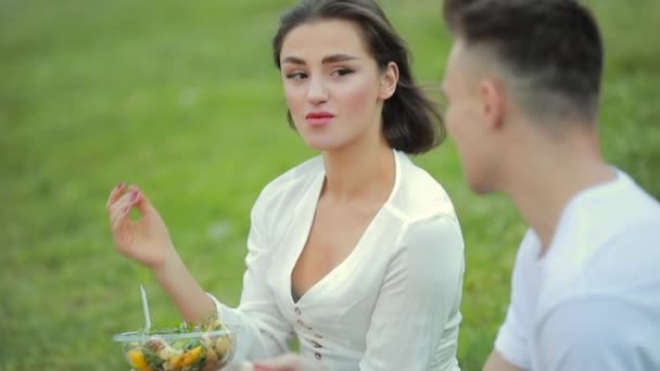 Pareja joven comiendo ensalada sentada en la hierba, picnic juntos — Vídeos de Stock