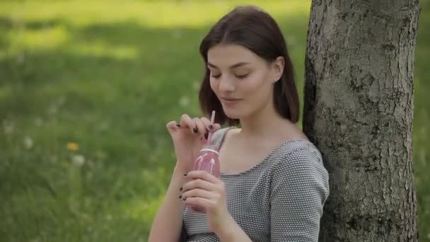 Jovem mulher positiva bebendo smoothie fresco através de uma palha sentada no parque de verão — Vídeo de Stock