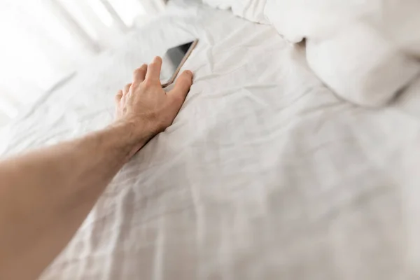 Мужская рука тянется к смартфону сразу после пробуждения в постели — стоковое фото