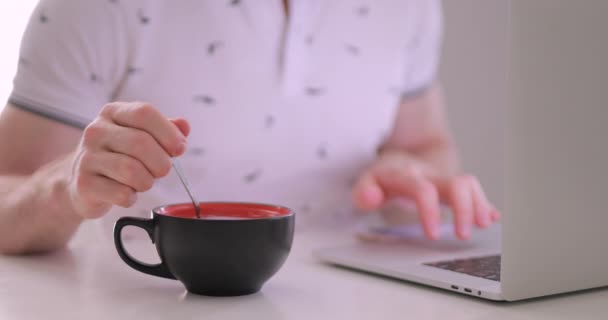 Primer plano del hombre mezclando azúcar en la taza de té mientras usa el ordenador portátil — Vídeos de Stock