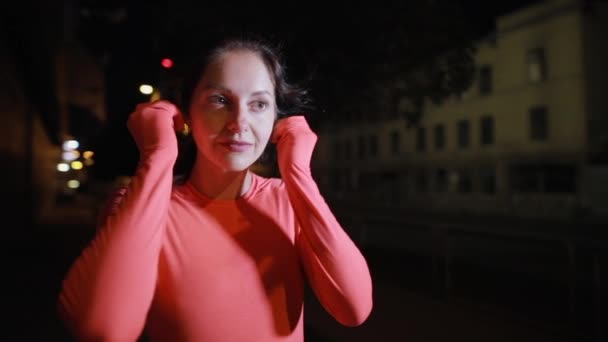 Donna sportiva che corre lungo la strada della città di notte si mette gli auricolari — Video Stock