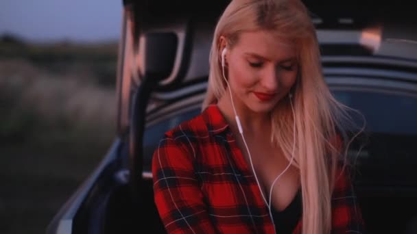 Sexy woman listening to music in earphones sitting on car — Stock Video