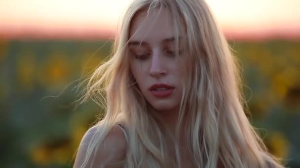 Женщина в поле на закате смотрит в камеру, ветер дует ее волосы — стоковое видео