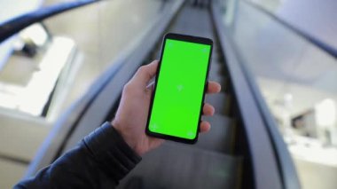 POV, alışveriş merkezinin yürüyen merdiveninde yeşil ekranlı akıllı telefon kullanan bir erkek.