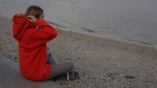 Vacker kvinna bär huva sitter vid havet stranden — Stockvideo