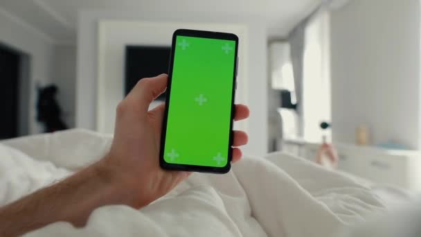 POV, tangan pria memegang smartphone dengan layar hijau di tempat tidur — Stok Video