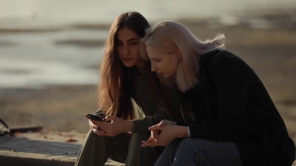 Due amiche sedute vicino al fiume e che guardano giù nello smartphone — Video Stock