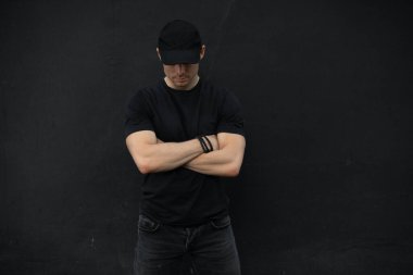 Siyah duvara yaslanmış kollarını kavuşturmuş, şapkalı, tanınmaz halde havalı bir adam.