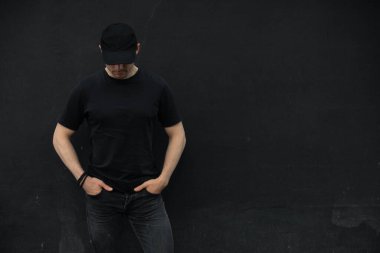 Siyah duvarın karşısında duran şapkalı ve boş tişörtlü tanınmayan bir adam.