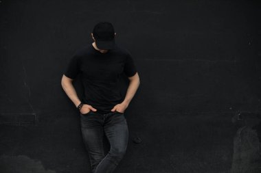 Siyah duvarın karşısında duran şapkalı ve boş tişörtlü tanınmayan bir adam.