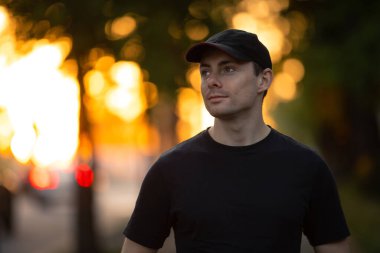 Siyah tişörtlü ve şapkalı şık bir adam yaz parkında yürüyor.