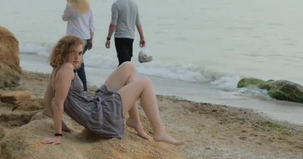 Кокетливая женщина в платье-дурачок, сидящая на морском пляже — стоковое видео