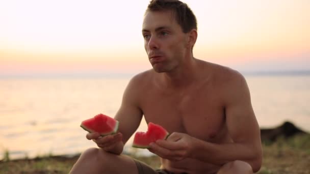 Человек ест арбуз сидя у моря — стоковое видео