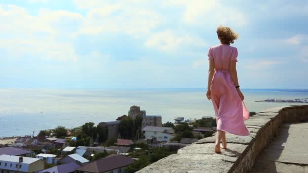 Mulher em um vestido rosa caminha ao longo do terraço contra o fundo do mar — Vídeo de Stock