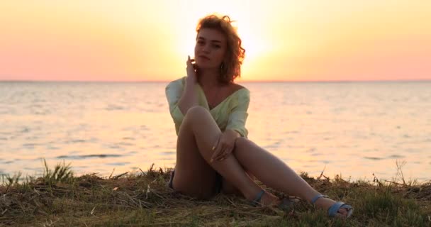 Όμορφη σέξι γυναίκα κάθεται κοντά στη θάλασσα στο ηλιοβασίλεμα — Αρχείο Βίντεο