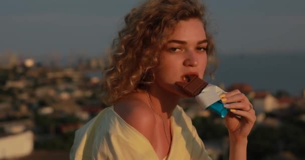 Кучерява жінка їсть шоколад над містом — стокове відео