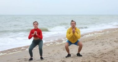 Okyanus sahilinde çömelme egzersizi yapan genç bir çift.