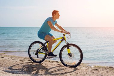 Genç adam deniz kıyısında bisiklet sürüyor.