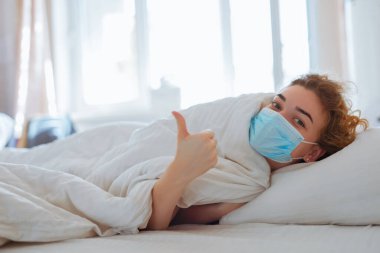 Cerrahi maskeli, koronavirüs koruması olan bir kadın yatakta yatıyor.