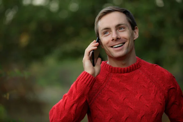 Man in red sweater talks by phone smiling outdoor at the fall day