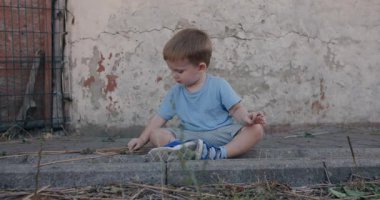 Little boy sitting outside near wall and playing with stick
