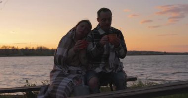 A man and a woman are sitting together on a bench by the river and drinking coffee