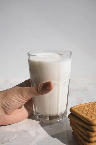 Mano sosteniendo un vaso lleno de leche en una mesa y un montón de galletas —  Fotos de Stock