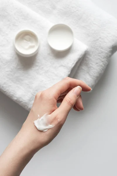 Close-up smooth moisturizer cream or body lotion on females hand in bathroom on white background with towel, indoors. Daily skincare routine vertical format, top view