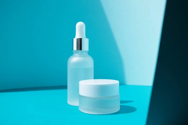 Buzlu cam şişe esaslı kolajen anti-yaşlanma serumu ve yüz nemlendirici kremi mavi kağıt arka planda, üst görünümde. Cilt bakımı güzellik ürünü