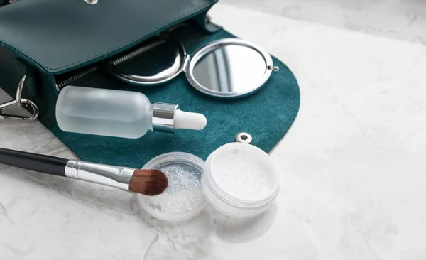 Mermer masanın üzerinde kozmetik ürünler olan küçük, şirin bir zümrüt kadın çantası. Şeffaf toz, makyaj fırçası, cep aynası, seçici odaklı yüz serumu. Güzelliği ve moda yaşam tarzını kavra