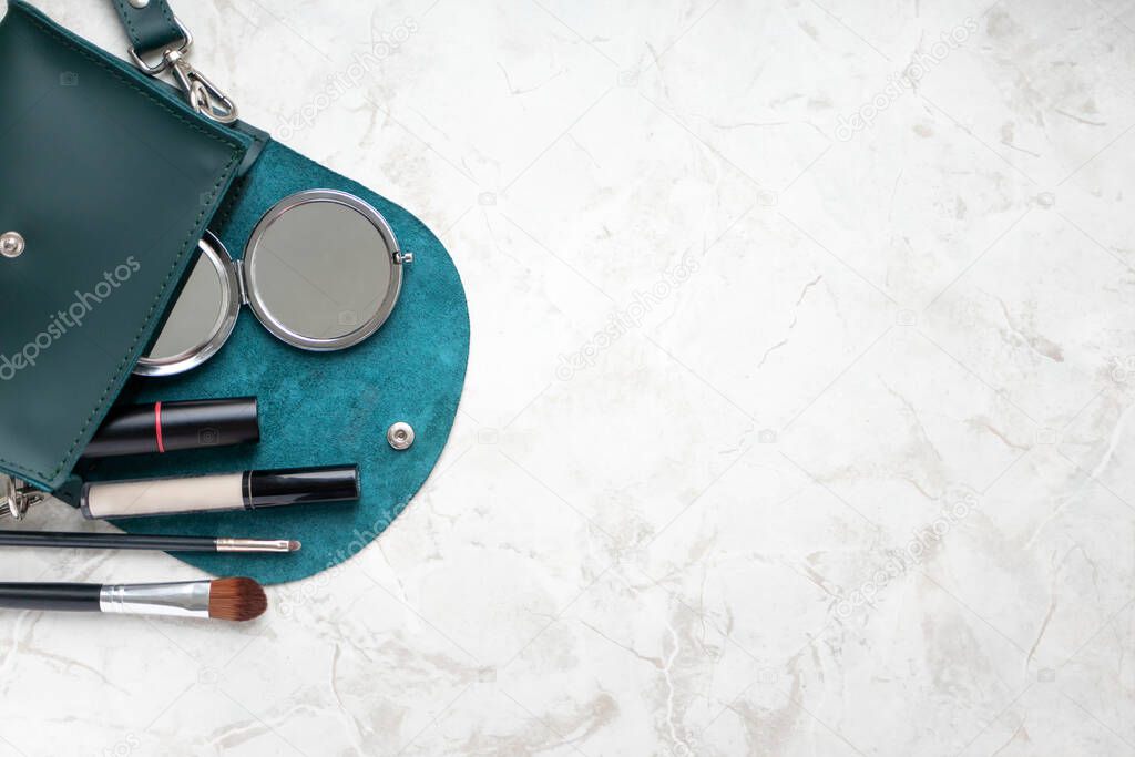 Compact pocket mirror, lipstick, concealer and makeup brushes in female bag on marble table with copy space top view. Beauty and fashion still life