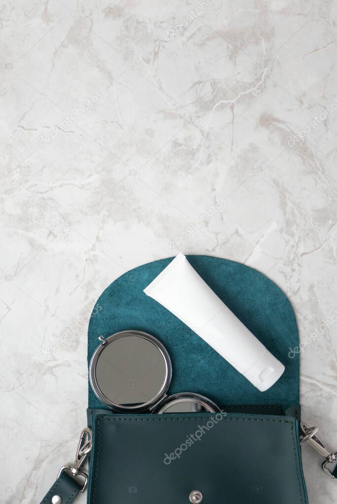 Moisturizer cream mockup and compact cosmetic mirror on emerald female bag in white marble background with copy space top view, vertical. Beauty product and accessory lifestyle flat lay. Concept for fashion blog