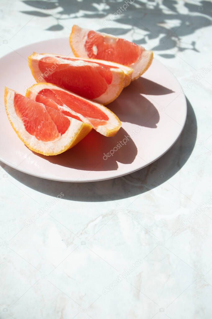 Cut grapefruit slices on pink plate on marble table with selective focus, copy space and trendy shadow, vertical. Fresh citrus rich source of antioxidants