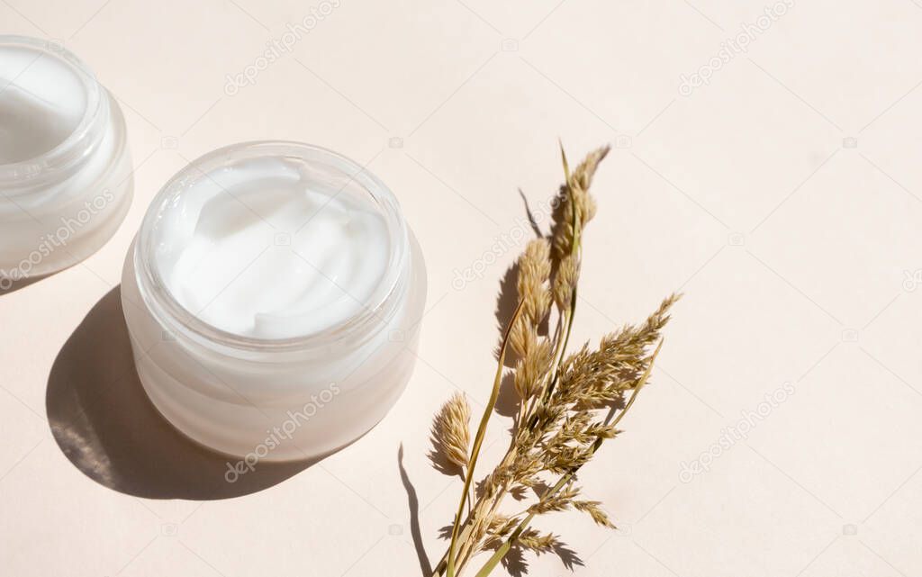 Moisturizer cream in open glass jar and dry meadow herbs on beige background, close-up. Care for sensitive skin with natural extract. Summer or autumn skincare body care concept