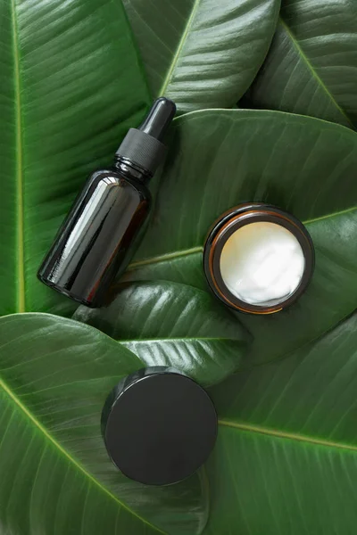 Facial hyaluronic serum fluid dropper and moisturizer cream in brown glass bottle and jar on freshness green tropical leaves background. Eco organic natural beauty skincare products. Dermatology and cosmetology