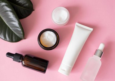 Açık nemlendirici kavanozlar, serum düşürücü cam şişeler, pembe arka planda el kremi ve yeşil tropikal yapraklar, üst manzara. Eko doğal organik özüt cilt bakım ürünleri bileşimi düzdü.