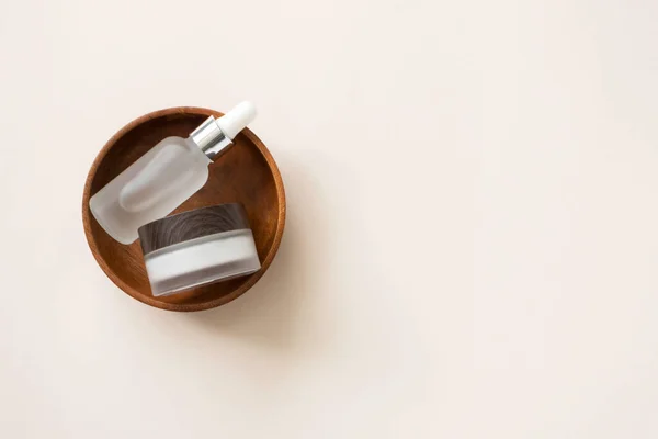 Cam şişedeki serum, pipetli ve yüz kremi nemlendiricili, ahşap tabakta, açık bej arka planda, fotokopi alanı, üst manzara. Minimalist cilt bakımı için doğal kozmetik ürünleri