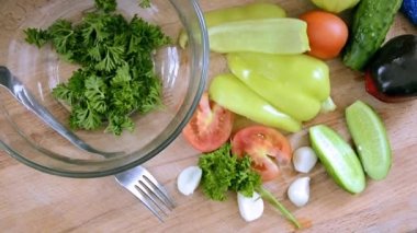 Sebzeler masada, salata dilimlemeye hazır. Bulgar biberi, domates, otlar, salatalık, sebze yemekleri için sarımsak. Kesme tahtasındaki cam kasenin yanında bir çatal ve bıçak..