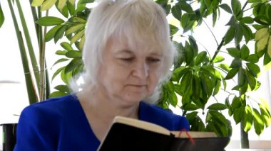 Yaşlı bir kadın kitap okur. Kadın, en sevdiği yazarın hikayelerini okurken durakladı. Kapalı alan bitkilerinin arka planında okuyan kadın.