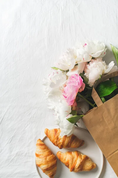 Bouquet of peonies in paper bag and croissants on white texture with copy space