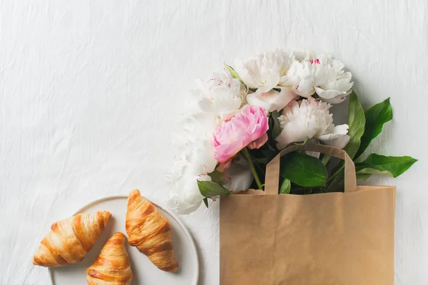 Bouquet of peonies in paper bag and croissants on white texture with copy space