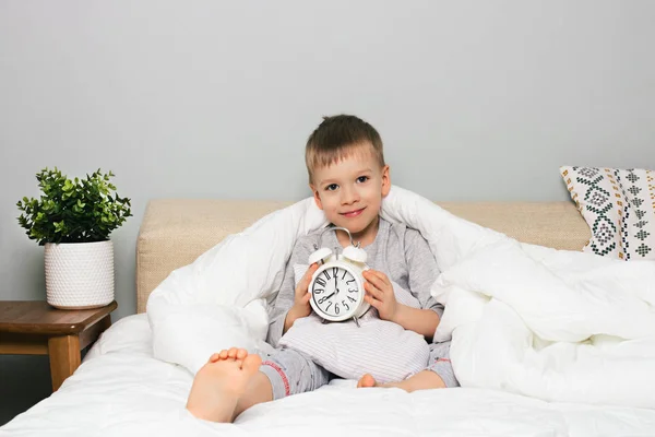 Cheerful little boy in bed with alarm clock on white background, wake up morning concept, sleeping time, copy space