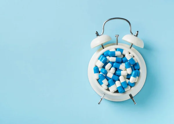 Heap of pills and capsules and alarm clock on blue background. Time and healthcare concept. Copy space, top view