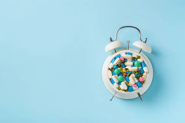Heap of colorful pills and capsules and alarm clock on blue background. Time and healthcare concept. Copy space, top view
