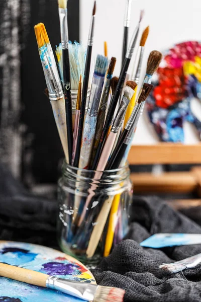 Pile of paint brushes in glass jar with palette and little easel on background