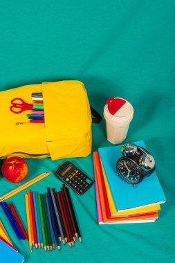 Sarı bir sırt çantası, defterler, renkli kalemler, bir elma, bir hesap makinesi ve bir çalar saatin yer aldığı turkuaz bir arka planda renkli okul malzemeleri.